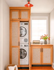 A laundry closet disguised as real furniture, with warm wood cabinetry and woven cane panel doors that fully conceal a stacked washer and dryer. A wood countertop, black hardware, and a styled vignette of art and tulips keep the space from feeling purely utilitarian. Overhead, a saturated orange flushmount with brass hardware adds the one bright, confident detail in an otherwise warm, neutral room.