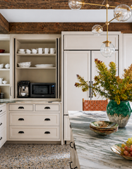 Cabildo brass globe chandelier with clear glass globe shades hanging above a rustic modern kitchen featuring white shaker cabinets, a dark reclaimed wood ceiling, and a large green floral arrangement, showcasing sculptural modern kitchen lighting.