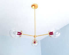 Deep Black Cherry and luminous brass give the Cadiz 3-light chandelier a rich, dramatic color story. This customizable Sazerac Stitches ceiling fixture features three clear glass globes, slender branching arms, and glossy wine-red powder-coated accents that emphasize its sculptural geometry. The combination offers distinctive lighting inspiration for moody dining rooms, jewel-toned bedrooms, dramatic entryways, and eclectic interiors mixing burgundy with warm metallic finishes. Explore statement globe light