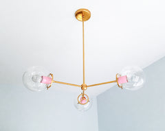 Blush Pink and Brass bring soft color to the Three-Light Cadiz Chandelier, a customizable modern globe light by Sazerac Stitches. Three clear glass globes extend from a slim branching brass frame, while pink powder-coated socket covers add a playful contrast to the warm metal. Its open, sculptural silhouette provides statement lighting without feeling visually heavy, making it an appealing chandelier for bedrooms, dining nooks, entryways, and rooms with lower visual clutter. Explore colorful contemporary li