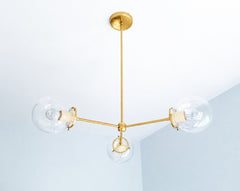 Cream and brass create a warm neutral palette on the Cadiz 3-light chandelier, a customizable modern ceiling light by Sazerac Stitches. Three clear glass globes branch from the slender brass frame, while glossy Cream powder-coated accents soften the sculptural design. This understated statement chandelier suits neutral dining rooms, serene bedrooms, elegant entryways, and contemporary interiors layered in ivory, beige, or warm white. Discover distinctive globe lighting with a quieter approach to color.