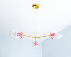 Vivid Doll Pink transforms the Cadiz 3-light chandelier into playful statement lighting for color-loving interiors. Designed by Sazerac Stitches, this customizable chandelier combines bright pink powder-coated details, warm brass arms, and three oversized clear glass globes in an airy sculptural silhouette. Consider it colorful ceiling-light inspiration for maximalist dining rooms, cheerful kitchens, creative studios, children’s rooms, or eclectic spaces with bold accent colors. Discover a modern globe chan