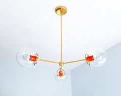 Flame orange and warm brass turn the Cadiz 3-light chandelier into bold statement lighting for colorful interiors. This customizable chandelier by Sazerac Stitches combines three clear glass globes with a streamlined branching brass frame and vivid Flame powder-coated accents. Its sculptural shape and saturated orange details offer striking inspiration for eclectic dining rooms, color-drenched kitchens, playful entryways, and contemporary spaces influenced by retro color palettes. Discover modern globe ligh