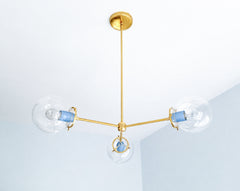 Hydrangea Blue and warm brass create an airy, elegant palette on the Cadiz 3-light chandelier by Sazerac Stitches. This customizable statement light features three clear glass globes, a slender branching brass frame, and glossy powder-coated blue accents. Its sculptural design offers fresh inspiration for blue dining rooms, colorful kitchens, serene bedrooms, traditional interiors with contemporary lighting, and eclectic spaces mixing pastel blue with metallic finishes. Discover modern globe lighting in a s