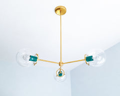 Mermaid Green and warm brass create a vibrant jewel-tone palette on the Cadiz 3-light chandelier by Sazerac Stitches. This customizable statement fixture pairs three clear glass globes with glossy green-blue powder-coated accents and a slender branching brass frame. Its airy, sculptural design offers colorful lighting inspiration for teal kitchens, eclectic dining rooms, dramatic entryways, and maximalist interiors combining turquoise, emerald, navy, or brass. Discover a modern globe chandelier in an expres