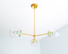 Mint green and brass give the Cadiz 3-light chandelier a fresh pastel palette with polished contemporary style. This customizable ceiling fixture by Sazerac Stitches combines three clear glass globes, glossy Mint powder-coated accents, and a slender branching brass frame. Its light, sculptural silhouette offers inspiration for pastel kitchens, cheerful breakfast rooms, colorful bedrooms, nurseries, and eclectic interiors mixing mint with blush, pale blue, cream, or warm metallic finishes. Discover distincti