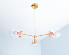 Peach and brass create a warm pastel color story on the Cadiz 3-light chandelier by Sazerac Stitches. Three clear glass globes extend from its slender branching frame, while glossy Peach powder-coated accents bring softness to the sculptural design. This customizable chandelier offers lighting inspiration for romantic bedrooms, colorful dining rooms, cheerful kitchens, nurseries, and eclectic interiors combining peach, blush, cream, or warm metallic finishes. Discover modern globe lighting with an inviting 