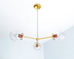 Matte Terra Cotta and warm brass bring an earthy, clay-toned palette to the Cadiz 3-light chandelier by Sazerac Stitches. This customizable statement fixture pairs three clear glass globes with a slender branching brass frame and muted reddish-brown powder-coated accents. Its sculptural design offers lighting inspiration for warm modern dining rooms, organic contemporary kitchens, bohemian bedrooms, and eclectic interiors layered with rust, cream, olive, ochre, or natural wood tones. Discover modern globe l