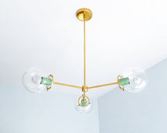 Soft Vista Green brings fresh color to the Cadiz 3-light chandelier, a customizable brass and glass fixture by Sazerac Stitches. Three clear glass globes extend from an airy branching frame, with glossy green powder-coated accents creating a gentle contrast against the warm brass. This sculptural chandelier offers lighting inspiration for green kitchens, botanical dining rooms, pastel bedrooms, serene entryways, and contemporary interiors with nature-inspired color palettes. Explore distinctive globe lighti