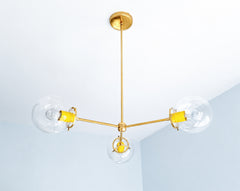 Sunny yellow accents bring joyful color to the Cadiz 3-light chandelier, a customizable brass and glass fixture by Sazerac Stitches. Three clear glass globes extend from its slender branching frame, while glossy yellow powder-coated details give the sculptural design a bright contemporary character. This colorful chandelier offers inspiration for cheerful kitchens, eclectic dining rooms, playful entryways, children’s spaces, and interiors mixing yellow with warm metallic finishes. Discover statement globe l