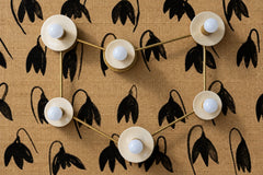 A cream and brass Capricorn constellation sconce with six globe bulbs on glossy cream discs, linked by brass rods that trace the stars of the sign. It sits on tan grasscloth wallpaper printed with loose black brushstroke flowers. Cream, black, and natural fiber give it a warm, graphic, organic modern look. Hang it over a bed, a sofa, or a dining banquette, or mount it as a ceiling light. A unique zodiac light for eclectic spaces and a great Capricorn gift.