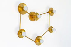 Brass constellation light fixture with six white globe bulbs, each set out from a round brass cup and linked by thin brass rods in an open star chart shape. From this angle you can see the round wall plate in the middle and how far the cups stand off the wall. Hang it as a large wall sconce over a bed or sofa, or as a flush mount ceiling light. Warm raw brass suits mid century modern and space age rooms, celestial bedroom lighting, and eclectic gold accented spaces.