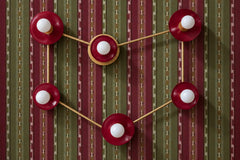 A burgundy and brass Capricorn constellation sconce with six globe bulbs on glossy Black Cherry discs, joined by brass rods that trace the stars of the sign. It hangs on a woven style stripe wallpaper in burgundy, olive green, and cream. Rich jewel tones and warm brass make it feel cozy and a little bohemian. Use it over a bed or sofa, in a den, or as a ceiling light. It suits maximalist decor, moody bedroom ideas, and unusual light fixtures for eclectic spaces.