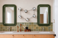 A nickel Capricorn constellation sconce with six globe bulbs on silver discs hangs centered between two puffy green framed mirrors. Below, a double vanity with a taupe quartz top, chrome faucets, light oak cabinets with round knobs, and a backsplash of mottled green square tile. Teak bud vases and a sculptural wood vase add warmth. It is retro and playful, a good fit for 70s inspired bathroom ideas, double vanity lighting, and unique sculptural wall sconces.