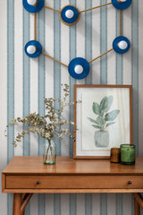A blue and brass Capricorn constellation light, its glossy disc shades and globe bulbs traced in brass like stars on a sky chart. It hangs over a walnut toned mid century console in an entry or home office. Blue and white zigzag stripe wallpaper, a glass jar of dried eucalyptus, a framed botanical print, and amber and green candles fill out the vignette. Fresh and a little retro, it suits entryway lighting, celestial decor, and unique wall sconce ideas.
