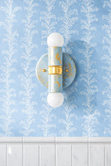 The Celestial, Cream, and Gold Marbled Small Thalia Sconce shown vertically on soft blue botanical wallpaper above glossy white wall tile. The handmade marbled finish gives this petite bathroom sconce a custom, small-batch feel, making it a lovely choice for powder rooms, kids bathrooms, coastal cottage spaces, grandmillennial decor, and soft blue wallpapered interiors.