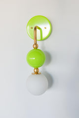 The Beaded Sconce in chartreuse stacks a hooked brass arm, small brass ball joints, and a glossy lime green glass sphere above a soft white opal glass globe, giving the whole fixture a bold, jewelry-like silhouette on the wall. It works well as a bedside light in place of a table lamp, beside a bathroom mirror, or anywhere a single statement sconce makes more sense than a matching pair.