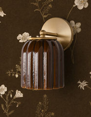 Cream wildflowers and delicate gold branches scatter across this deep espresso wallpaper, and the Marcella Sconce in Chicory Brown matches the dark ground so closely that the flowers seem to grow right out of the fixture, with only the brass hardware marking where wallpaper ends and sconce begins. A moody, botanical pairing for dark academia bedrooms and rooms that want floral wallpaper without going pale or sweet.
