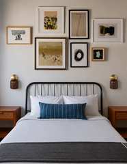 A confidently mismatched gallery wall hangs above this bedroom's black iron bed frame, mixing gold, black, and white frames around botanical prints, abstract art, and a soft landscape. A symmetrical pair of Marcella Sconces in Chicory Brown grounds the arrangement at bedside height, their glossy amber shades and brass hardware adding warmth beneath all that curated art. Gallery wall bedroom inspiration for anyone who wants collected, not matched.