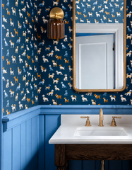 Dozens of illustrated terriers and hounds scatter across navy wallpaper in this powder room, paired with periwinkle wainscoting and a brass-framed mirror for a look that's polished but never stuffy. The Marcella Sconce in Chicory Brown picks up the wallpaper's warm accent tones, its fluted glossy shade glowing like an old lodge lantern beside the vanity. A confident, collected take on dog wallpaper bathroom ideas for anyone who wants pattern with a sense of humor.