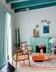 Chrome Aries constellation wall sconce with four sculptural metal disc shades and white globe bulbs, styled above a teal tile fireplace in an airy coastal boho living room. Soft seafoam walls with teal ceiling beams, sheer aqua curtains, tall cactus plants, midcentury wood lounge chairs with woven seats, and a round modern rug with a coral edge create a relaxed beach house vibe. Eclectic, colorful interior design with a bright orange lacquer coffee table and palm tree wall art, perfect statement lighting 