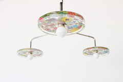 Close-up of a modern ceiling light fixture with three circular lampshades embedded with colorful pressed flowers, creating a bright and artistic design against a white background.