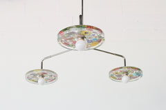 Close-up of a modern ceiling light fixture with three circular lampshades embedded with colorful & rainbow pressed flowers & greenery, creating a bright and artistic design against a white background.