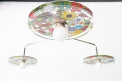 Close-up of a modern ceiling light fixture with three circular lampshades embedded with colorful pressed flowers, creating a bright and artistic design against a white background.