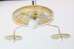 Close-up of a modern ceiling light fixture with three circular lampshades embedded with pressed yellow flowers and greenery, creating a bright and artistic design against a white background.