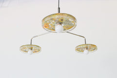 Close-up of a modern ceiling light fixture with three circular lampshades embedded with yellow flowers, pressed leaves, and greenery, creating a bright and artistic design against a white background.