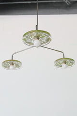 Close-up of a modern ceiling light fixture with three circular lampshades embedded with pressed leaves and greenery, creating a bright and artistic design against a white background.