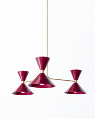 Deep black cherry and warm brass is the pairing that makes a room feel rich, sultry, and a little glamorous, and this mid-century modern Clancy chandelier brings them together beautifully. Three glossy magenta-burgundy double-cone shades radiate from a brass central hub on slim brass arms, in a fun, sculptural MCM silhouette that throws light up and down from each arm. The brass stem and collars warm up the saturated jewel tone and give the whole piece a polished, collected feel. It's built to be the star o