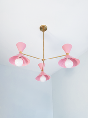 Soft blush pink and warm brass is the pairing that makes a room feel sweet, romantic, and a little playful, and this mid-century modern Clancy chandelier brings them together beautifully. Three glossy blush double-cone shades radiate from a brass central hub on slim brass arms, in a fun, sculptural MCM silhouette that throws light up and down from each arm. Seen from below, you can read the whole graphic layout, brass stem, brass canopy, and that soft pink glow. It's built to be the star over a dining table