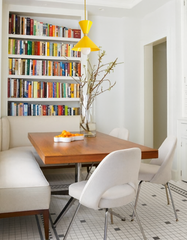 Contemporary breakfast nook featuring a bold yellow and brass Clancy pendant light above a square wood dining table with cream upholstered banquette seating and modern curved dining chairs. The bright space has white tiled floors, a built-in bookshelf filled with colorful books, and a vase with branches, blending mid-century lighting with modern eclectic design.