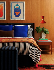Pair of Flame red Clancy wall sconces with sleek brass accents warm up a midcentury-inspired bedroom. Mounted beside a bed styled with cobalt blue pillows, striped bedding, and an orange throw, these bold wall lights from Sazerac Stitches show how vibrant, customizable lighting can add personality to any modern space.