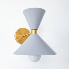 A calm gray wall sconce that brings quiet mid-century character to a powder room, hallway, or bedside. The Clancy Sconce carries the same sculptural double-cone shade as the Clancy chandelier, a wide cone below and a smaller cone flaring up above, joined at a raw brass collar and mounted on a round brass backplate. Soft gray reads neutral without going flat, the kind of easy color that works against white trim, wallpaper, tile, or a moodier wall. A versatile light for mid-century, transitional, or colorful 