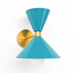 A cheerful blue-teal wall sconce that brings easy coastal color to a powder room, bedside, or hallway. The Clancy Sconce carries the same sculptural double-cone shade as the Clancy chandelier, a wide cone below and a smaller cone flaring up above, joined at a raw brass collar and mounted on a round brass backplate. This soft dusty teal reads fresh and a little coastal, lovely against white trim, patterned tile, natural wood, or a crisp wallpaper. A friendly hit of color for mid-century, coastal, or colorful