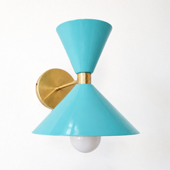 A breezy poolside-aqua wall sconce that brings happy, retro color to a powder room, bathroom, or kids' space. The Clancy Sconce carries the same sculptural double-cone shade as the Clancy chandelier, a wide cone below and a smaller cone flaring up above, joined at a raw brass collar and mounted on a round brass backplate. This light aqua reads cheerful and a little Palm Springs, lovely against white subway tile, rattan, natural wood, or a crisp wallpaper. A joyful hit of color for coastal, mid-century, or c