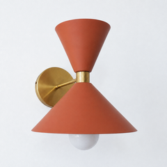 A warm terracotta wall sconce that brings earthy, sun-baked color to a powder room, bedside, or hallway. The Clancy Sconce carries the same sculptural double-cone shade as the Clancy chandelier, a wide cone below and a smaller cone flaring up above, joined at a raw brass collar and mounted on a round brass backplate. This matte clay tone reads grounded and organic, beautiful against white plaster, natural wood, zellige tile, or a moody plaster wall. A warm, characterful pick for mid-century, organic modern,