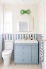 This photo shows a fresh, airy bathroom with a soft coastal feel, featuring a mint green Coquille double sconce mounted above a simple mirror. Pale blue striped wall tile, a powder blue vanity, warm brass hardware, and creamy white walls give the space a light, polished look that feels both classic and playful. The shell-inspired sconce adds a sculptural focal point and brings in a subtle beachy influence without looking themed or cheesy. This image would appeal to anyone searching for coastal bathroom ligh