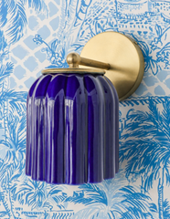 Layered palm fronds and a subtle lattice print cover this wallpaper in classic blue and white, and the Marcella Sconce in Cobalt Blue pulls its exact color straight from the pattern. The glossy fluted shade and brass ball finial make the fixture feel like part of the wallpaper's story rather than a separate choice. Preppy coastal inspiration for bathrooms, powder rooms, and bedrooms that want blue and white with real depth.