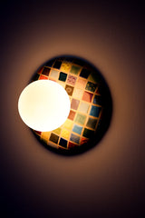 Colorful Cube Glazed Globe Sconce installed on wall and lit, handmade ceramic wall sconce with glossy multicolor cube grid glaze in reds, blues, greens, yellows, and neutrals, paired with a matte white glass globe shade, warm ambient light and soft glow, statement wall lighting for maximalist interiors, modern eclectic home decor, playful bedside sconces, bold hallway lighting, and colorful accent lighting for design forward spaces