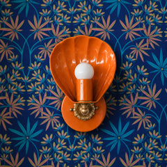 Bright orange against deep cobalt-blue tropical wallpaper — the kind of high-contrast color pairing that makes a maximalist room sing. The Coquille sconce from Sazerac Stitches is a glossy scallop-shell wall light with a warm brass fitting and scalloped floral backplate, here in a saturated orange-peel hue set against a botanical palm-leaf print in pink, teal, and chartreuse. A statement lighting moment for a color-drenched powder room, an eclectic bedroom, or a bold entryway. Works for maximalist decor, co