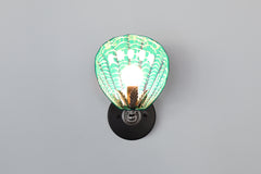 Coquille Sconce lit up, showing vibrant mermaid green ceramic shell with glowing painted patterns and silver-toned leaf detail around the bulb; mounted on a black wall plate, handmade and one of a kind