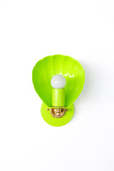 A sculptural chartreuse shell wall sconce shown against a white background, featuring a glossy scallop-inspired shade, matching chartreuse backplate and socket cover, a white globe bulb, and warm brass detailing with an ornate floral rosette accent. The Coquille sconce blends playful color with a refined coastal feel, making it ideal for designers searching for chartreuse lighting, shell sconces, tropical bathroom lighting, luxury pool bathroom sconces, boutique hotel lighting, Palm Beach style interiors, B