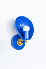 Angled studio photo of the Mediterranean Blue Coquille Sconce, a glossy blue shell-shaped wall light with a round backplate, white globe bulb, and warm brass rosette detail. The scalloped shade gives it a coastal, Art Deco, and vintage-inspired look while the saturated blue keeps it playful and bold. A strong fit for bathroom vanity lighting, powder rooms, blue and white interiors, nautical decor, beach house lighting, kids bathrooms, bedside sconces, and colorful hallway lighting.