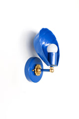 Angled studio photo of the Mediterranean Blue Coquille Sconce, a glossy blue shell-shaped wall light with a round backplate, white globe bulb, and warm brass rosette detail. The scalloped shade gives it a coastal, Art Deco, and vintage-inspired look while the saturated blue keeps it playful and bold. A strong fit for bathroom vanity lighting, powder rooms, blue and white interiors, nautical decor, beach house lighting, kids bathrooms, bedside sconces, and colorful hallway lighting.