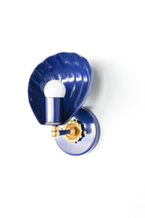 Angled studio photo of a navy shell wall sconce with a sculptural scallop shade, brass rosette detail, and white globe bulb. The Coquille Sconce blends coastal lighting, Art Deco curves, and colorful modern hardware in a compact shape that can bring personality to small spaces. Use this kind of navy and brass wall light for bathroom vanity lighting, a powder room, a bedside reading light, a hallway accent, or a blue and white coastal interior.