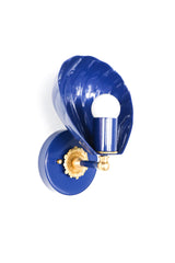Angled side view of the Navy Coquille Sconce showing the depth of the glossy shell shade, the round navy wall plate, brass hardware, and white globe bulb. The scalloped silhouette makes this a decorative wall sconce for anyone looking for navy bathroom lighting, shell sconces, coastal powder room ideas, nautical wall lights, or a small blue sconce with brass details. It has a collected, boutique hotel feel that would work in a bathroom, bedroom, hallway, or entry.