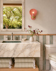 Coastal powder room styling featuring a peach Coquille shell wall sconce with warm brass details, mounted above a marble vanity and sink. Soft seaside materials include vertical seafoam tile wainscoting, an oversized stone-framed mirror, and a cushioned striped bench with fringe. Fresh flowers and collected decor add a relaxed, elevated look. Ideal inspiration for coastal bathroom lighting, scallop shell sconce styling, pink wall sconce, brass bathroom sconces, nautical cottage, and grandmillennial interior